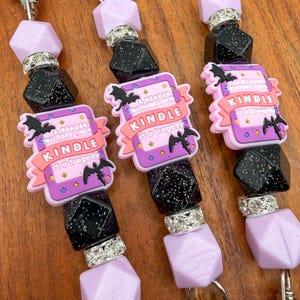 May include: Three beaded keychains with silver clasps. Each keychain features a pink and purple Kindle charm with the text "A reader without their Kindle is a tragedy." The beads are black, silver, and lavender.