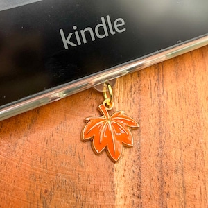 May include: A small, bright orange maple leaf charm with a gold-toned metal outline and a matching gold jump ring. The charm is attached to the bottom edge of a black electronic device with the word kindle printed in white on the screen.