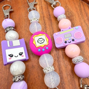 May include: Keychains with retro-themed charms. The keychains feature a purple floppy disk, a pink Tamagotchi, and a purple boombox. The charms are connected with beaded strands of purple, white, and clear beads, accented with silver rhinestone spacers. Each keychain has a silver clasp.