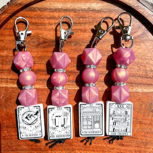 May include: Four keychains with silver clasps, featuring pink beads and tarot card-style charms. The charms display black and white illustrations and text, including "The Reader," "The Audiobook," "The Bookshop," and "The Tropes."