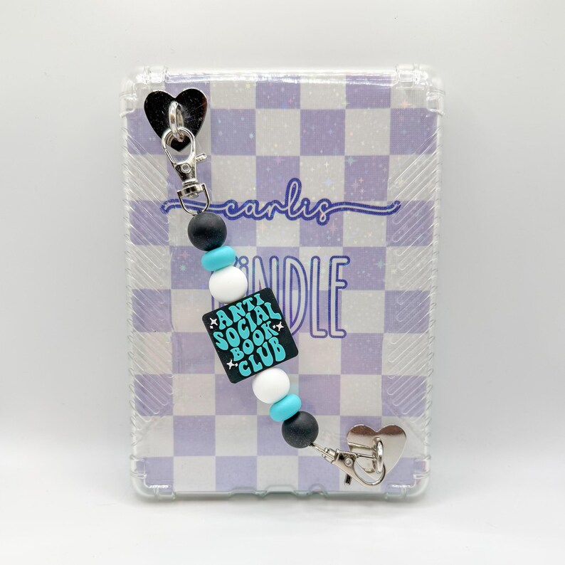 May include: A clear case with a purple and white checkered pattern, featuring a beaded charm. The charm has black, white, and turquoise beads, and a black square with the text "ANTI SOCIAL BOOK CLUB". The charm has a silver heart-shaped clip.