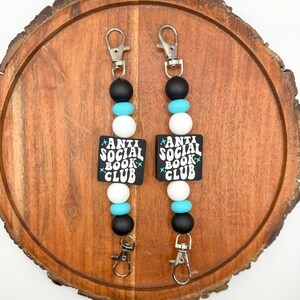 May include: Two keychains with black, white, and turquoise beads. Each keychain has a square black charm with the words "ANTI SOCIAL BOOK CLUB" in white. The keychains have silver-colored clasps.