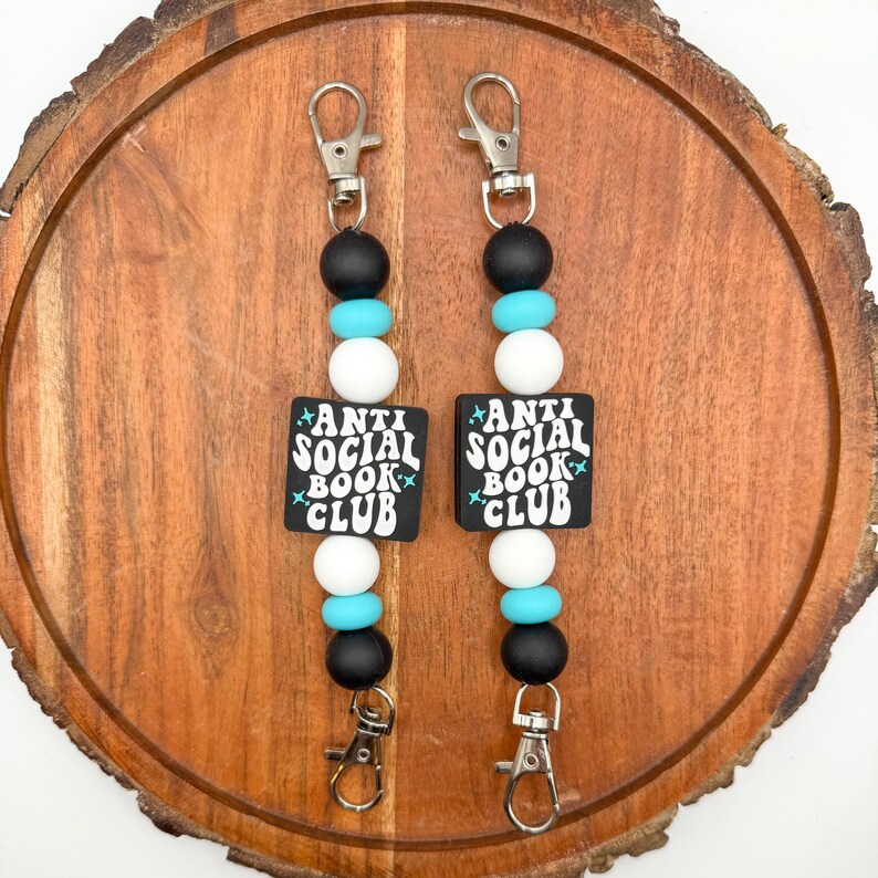 May include: Two keychains with black, white, and turquoise beads. Each keychain has a square black charm with the words "ANTI SOCIAL BOOK CLUB" in white. The keychains have silver-colored clasps.
