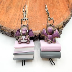 May include: Two keychains featuring purple glittery dragon figurines with outstretched wings. Each dragon sits atop a stack of three miniature books in pastel colors, attached to a silver clasp.