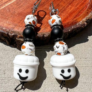 May include: A pair of Halloween-themed earrings. Each earring features a white chef hat with a smiling face, connected to a strand of beads. The beads include black spheres and white beads with pumpkin and skull patterns. Silver-tone ear hooks.