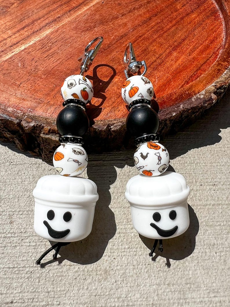 May include: A pair of Halloween-themed earrings. Each earring features a white chef hat with a smiling face, connected to a strand of beads. The beads include black spheres and white beads with pumpkin and skull patterns. Silver-tone ear hooks.