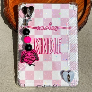 Op de afbeelding: Transparante Kindle-hoes met een roze en wit geblokt patroon. De hoes heeft een bedel met zwarte en roze kralen en een roze bedel met de tekst "You had me at Trigger Warnings". Het woord "Kindle" is roze.