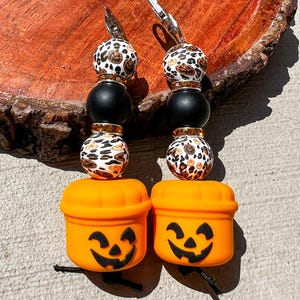 May include: Two Halloween-themed beaded keychains. Each keychain features a pumpkin bucket charm with a carved face, black beads, and leopard print beads. The keychains have silver-colored metal clasps.