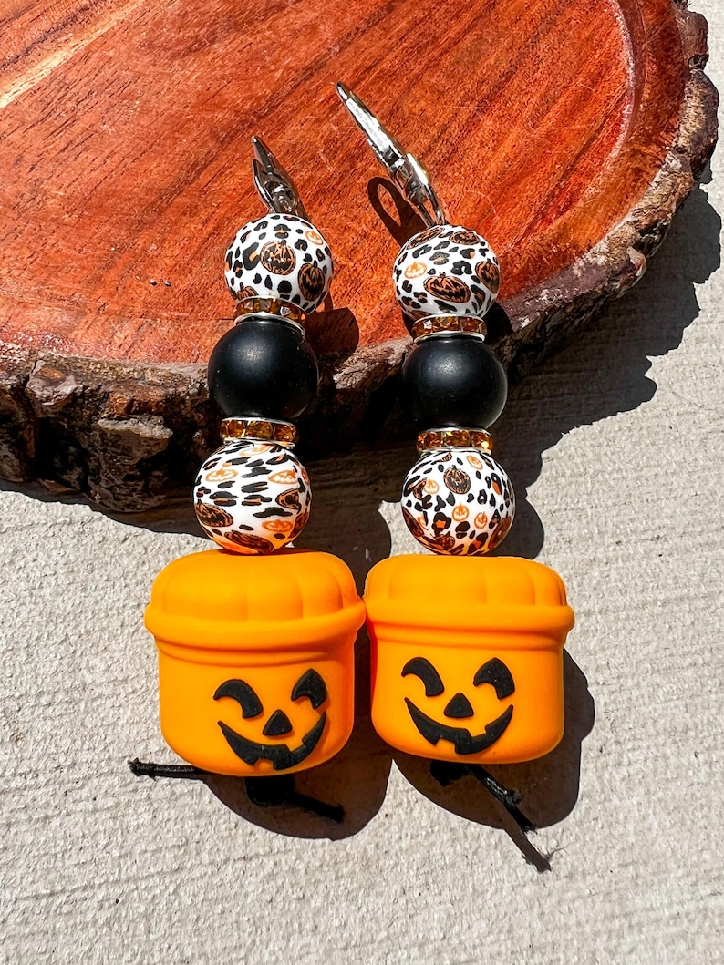 May include: Two Halloween-themed beaded keychains. Each keychain features a pumpkin bucket charm with a carved face, black beads, and leopard print beads. The keychains have silver-colored metal clasps.