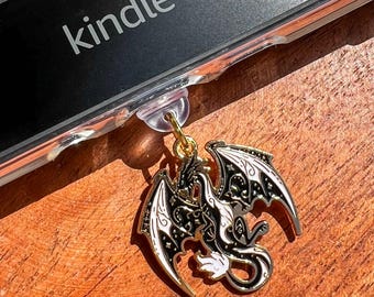 May include: A gold-colored dragon charm with black and white details hangs from a clear phone case. The word "kindle" is visible on the phone case. The charm is on a wooden surface.