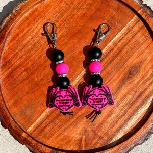 Op de afbeelding: Twee sleutelhangers met zwarte en roze kralen en zilveren sluitingen. Elke sleutelhanger heeft een roze bedel met de tekst "You had me at Trigger Warnings" en een skelet hand ontwerp. Op een houten oppervlak.
