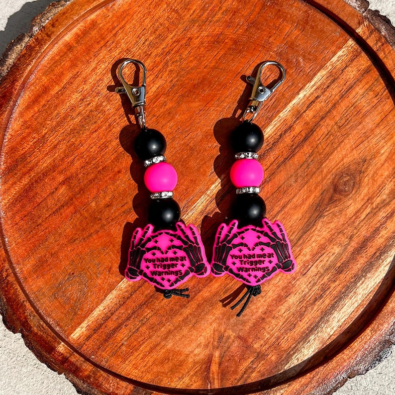 Op de afbeelding: Twee sleutelhangers met zwarte en roze kralen en zilveren sluitingen. Elke sleutelhanger heeft een roze bedel met de tekst "You had me at Trigger Warnings" en een skelet hand ontwerp. Op een houten oppervlak.