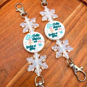 May include: Two identical badge reels featuring a white circular center with the text Chilly Nights and Books of Spice, decorated with small icons of handcuffs, a mug, and a book. Each reel has two iridescent snowflake charms and silver clips.