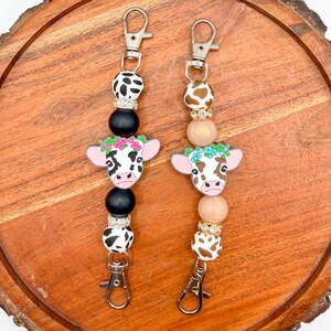 May include: Two beaded lanyards featuring silver-tone metal lobster clasps at both ends. One lanyard has black and white cow-print beads with a cow charm wearing a pink flower crown. The other has brown and white cow-print beads with a cow charm wearing a blue flower crown.