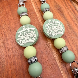 May include: Two beaded lanyards featuring sage green silicone beads, silver-toned rhinestone spacers, and a round sage green focal bead with white text that reads it's too nonfiction outside above an illustration of an open book with flowers.