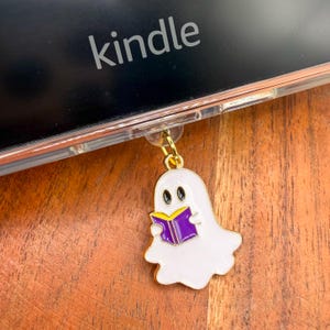 May include: A white ghost-shaped enamel charm with black eyes, holding an open purple book with yellow pages. The charm features a gold-toned metal jump ring attached to the clear plastic case of a Kindle e-reader with the word kindle visible.