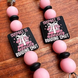 May include: Two pink and black beaded lanyards with silver-tone clasps. Each lanyard features a black square with the text "SUSPECT IS definitely READING SMUT" in white and pink, along with a graphic of a stack of books and a bow.