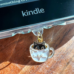 May include: A gold charm with a black cat peeking out of a white coffee cup. The charm is attached to a black Kindle e-reader with the word "kindle" printed on the front.