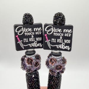 May include: Two black pens with crystal embellishments and decorative tops. Each pen has a black square with the text "Give me touch her & I'll kill you vibes" in white, a pink heart, and a dagger illustration.