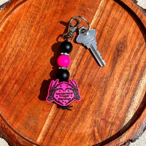 Op de afbeelding: Een sleutelhanger met een zilveren sluiting, een sleutel en een kralen bedel. De bedel heeft zwarte en roze kralen en een roze label met de tekst "You had me at Trigger Warnings" en een skelet hand ontwerp.