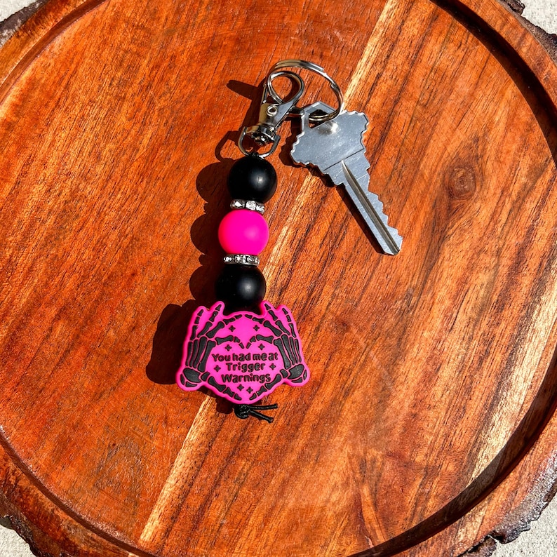 Op de afbeelding: Een sleutelhanger met een zilveren sluiting, een sleutel en een kralen bedel. De bedel heeft zwarte en roze kralen en een roze label met de tekst "You had me at Trigger Warnings" en een skelet hand ontwerp.