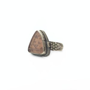 Puede incluir: Un anillo de plata con una piedra triangular de color marrón claro. La piedra tiene texto descolorido. La banda del anillo tiene un diseño texturizado. El engaste alrededor de la piedra tiene un diseño similar a una cuerda.