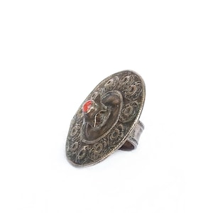 May include: A silver ring with a large, round, ornate top featuring a red stone in the center. The ring is designed with intricate floral patterns.