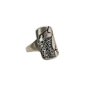 Könnte beinhalten: Ein silberfarbener Ring mit einer breiten, rechteckigen Vorderseite und einer strukturierten Oberfläche. Das Design umfasst einen Bereich mit kleinen, unregelmäßigen Vertiefungen und dunklen, geätzten astähnlichen Linien.