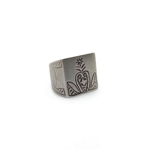 May include: A silver-colored, square ring with engraved floral and geometric designs. The ring features a central floral motif with heart-shaped leaves and a flower at the top. The ring is set against a white background.