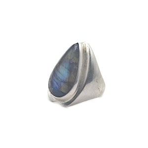 May include: A silver ring featuring a large, teardrop-shaped labradorite stone. The stone displays iridescent blue and green hues. The ring has a wide, smooth band and a bezel setting that securely holds the stone.