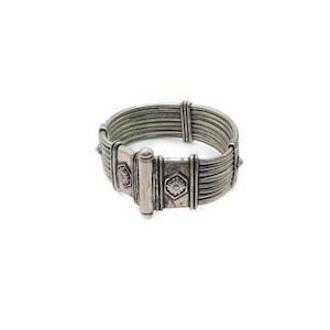 May include: A silver-toned cuff bracelet featuring multiple parallel wire strands held together by vertical metal bars. The central section includes two rectangular plates, each embossed with a small floral design within a hexagonal border.