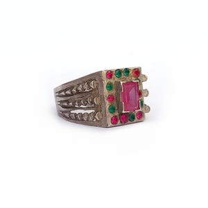 May include: A silver ring featuring a central rectangular pink gemstone, framed by smaller red and green stones. The ring's band has a textured pattern. The design is set against a white background.