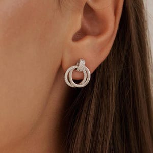 Puede incluir: Pendientes de plata con un diseño de doble círculo, cada círculo está incrustado con pequeñas piedras transparentes.