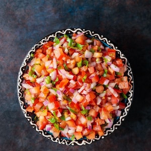 May include: A vibrant bowl of fresh salsa, featuring diced red tomatoes, red onion, green peppers, and cilantro. The salsa is served in a black bowl with a decorative white and black rim, set against a dark blue background.
