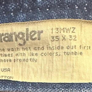 Pode incluir: Close-up de uma etiqueta de jeans Wrangler. A etiqueta &eacute; bege com texto preto, incluindo o nome da marca, o tamanho 35 x 32 polegadas (89 x 81 cm) e as instru&ccedil;&otilde;es de lavagem. A etiqueta diz "Made in USA" e "100% Algod&atilde;o".