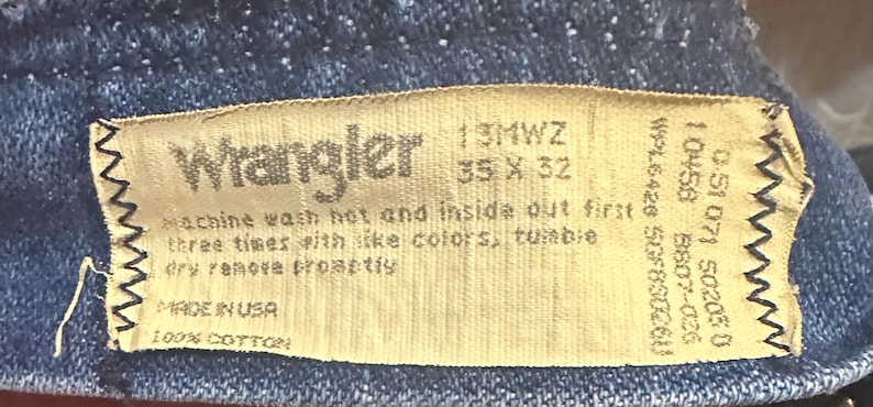 Pode incluir: Close-up de uma etiqueta de jeans Wrangler. A etiqueta &eacute; bege com texto preto, incluindo o nome da marca, o tamanho 35 x 32 polegadas (89 x 81 cm) e as instru&ccedil;&otilde;es de lavagem. A etiqueta diz "Made in USA" e "100% Algod&atilde;o".