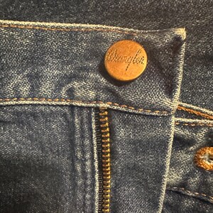 Pode incluir: Close-up de jeans azuis com um bot&atilde;o e z&iacute;per de cor lat&atilde;o. O bot&atilde;o &eacute; gravado com o nome da marca "Wrangler". Os jeans t&ecirc;m costuras vis&iacute;veis e um design cl&aacute;ssico.