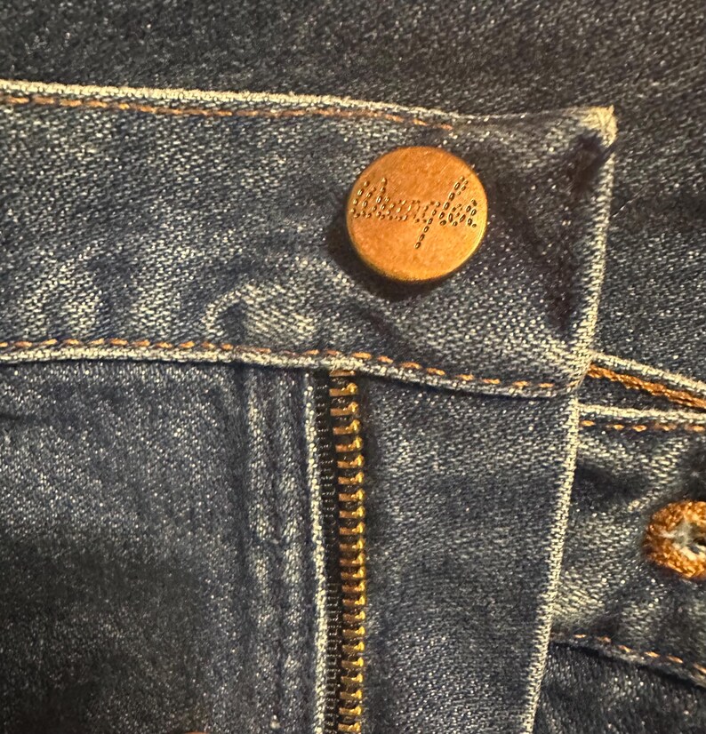 Pode incluir: Close-up de jeans azuis com um bot&atilde;o e z&iacute;per de cor lat&atilde;o. O bot&atilde;o &eacute; gravado com o nome da marca "Wrangler". Os jeans t&ecirc;m costuras vis&iacute;veis e um design cl&aacute;ssico.