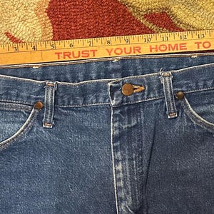 Pode incluir: Close-up de cal&ccedil;as de ganga azuis com um design cl&aacute;ssico de cinco bolsos. As cal&ccedil;as de ganga t&ecirc;m fecho de bot&atilde;o e z&iacute;per, e os bolsos s&atilde;o refor&ccedil;ados com rebites. Uma r&eacute;gua de madeira com o texto "TRUST YOUR HOME TO SUPERMIX" &eacute; colocada sobre as cal&ccedil;as de ganga.