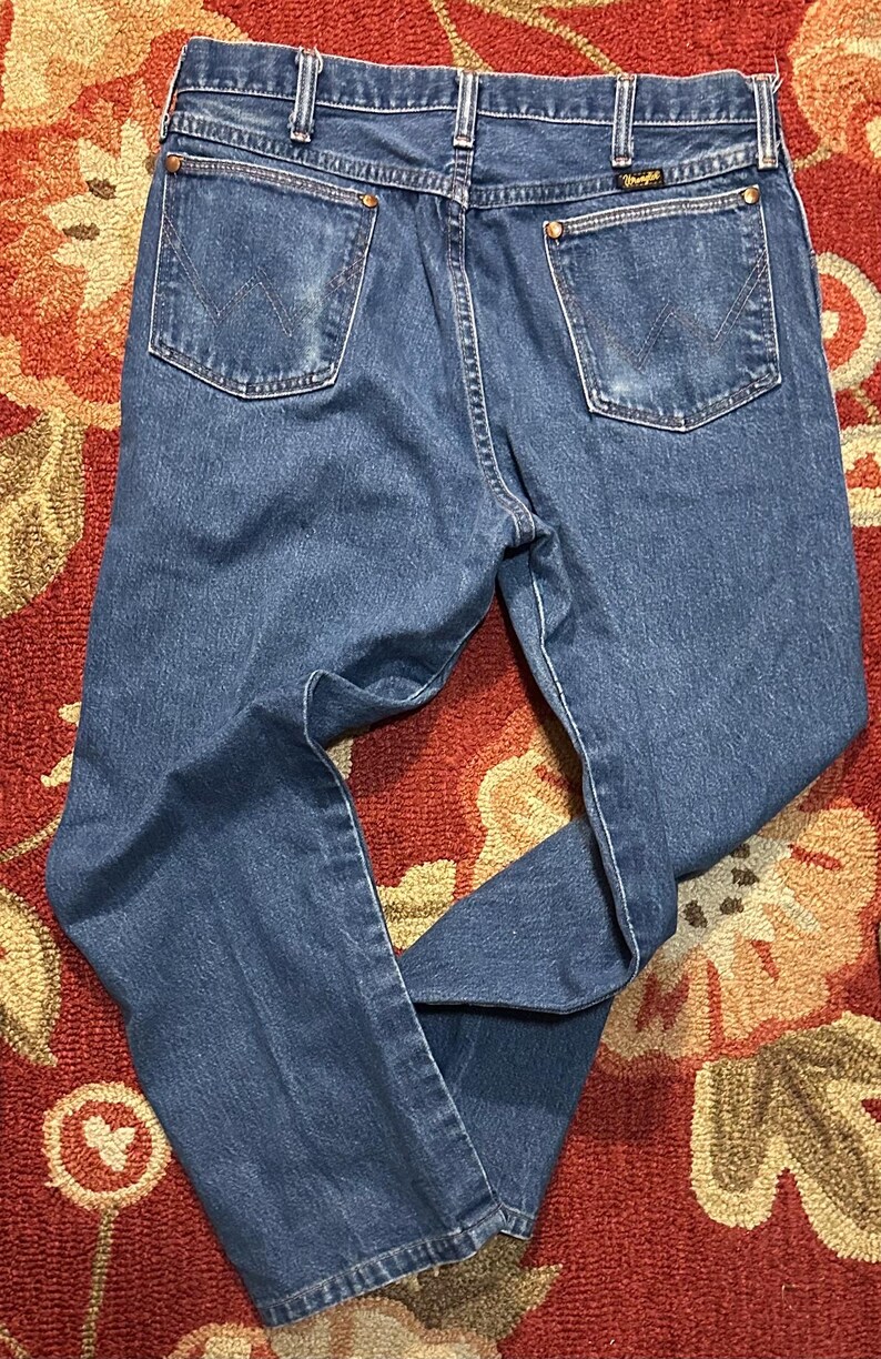 Pode incluir: Cal&ccedil;as de ganga azuis com um design cl&aacute;ssico de cinco bolsos. As cal&ccedil;as de ganga apresentam um corte de perna reto, presilhas para cinto e o log&oacute;tipo Wrangler no bolso traseiro. As cal&ccedil;as de ganga s&atilde;o de lavagem m&eacute;dia e mostram algum desgaste.