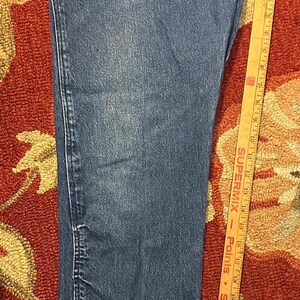 Pode incluir: Close-up de jeans azuis. Os jeans t&ecirc;m um design cl&aacute;ssico de cinco bolsos com perna reta. Uma r&eacute;gua de madeira com medidas em polegadas est&aacute; ao lado dos jeans.