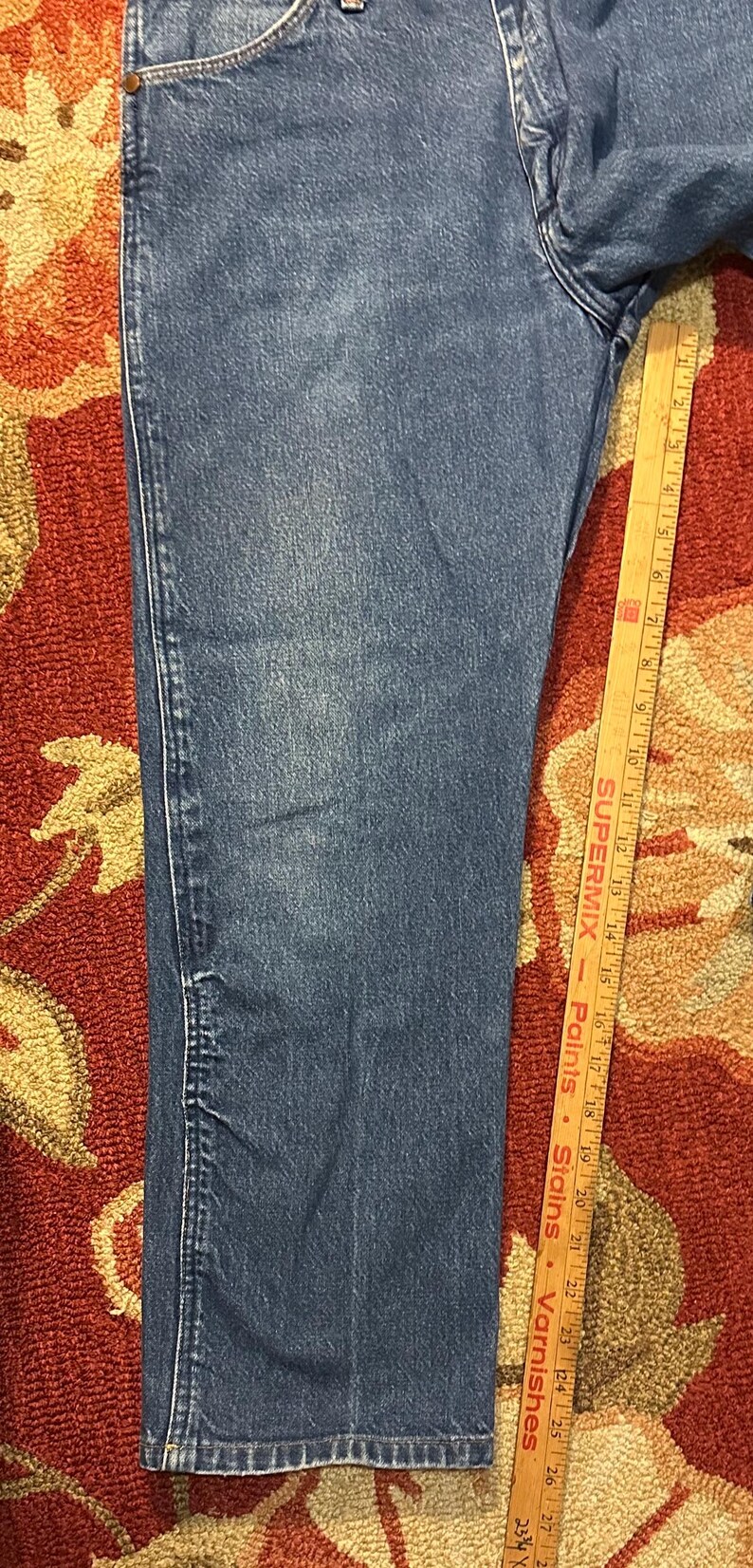 Pode incluir: Close-up de jeans azuis. Os jeans t&ecirc;m um design cl&aacute;ssico de cinco bolsos com perna reta. Uma r&eacute;gua de madeira com medidas em polegadas est&aacute; ao lado dos jeans.