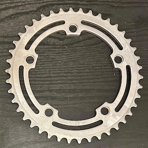 Puede incluir: Un plato de cadena de bicicleta de plata con un conteo de 42 dientes. El plato tiene una forma circular con un agujero central y una serie de dientes alrededor del borde exterior. El plato está diseñado para ser utilizado con una cadena de bicicleta.