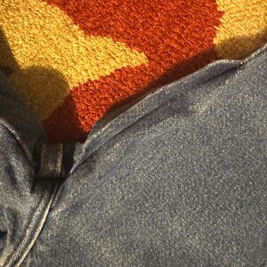 Pode incluir: Close-up de jeans de ganga azul. Os jeans est&atilde;o ligeiramente enrugados, com costuras vis&iacute;veis e fecho de bot&atilde;o. O tecido tem um aspeto texturizado. O fundo apresenta um tapete colorido com tons de laranja, amarelo e castanho.