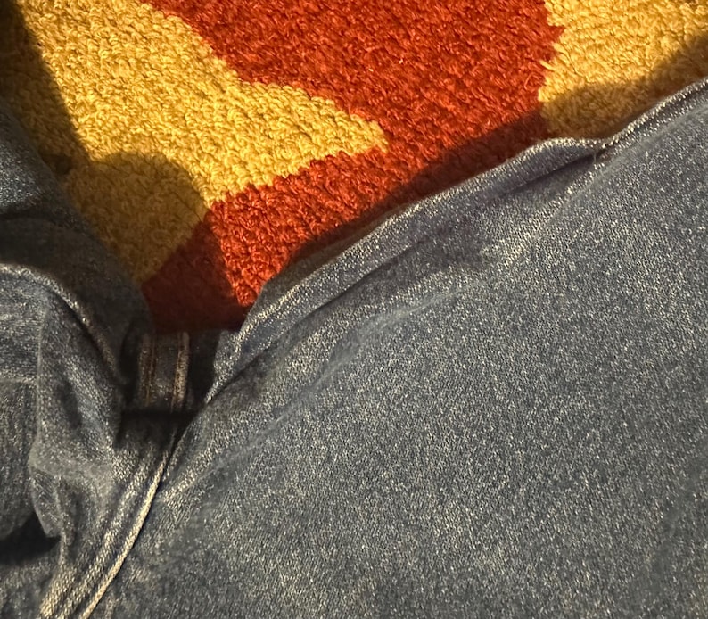 Pode incluir: Close-up de jeans de ganga azul. Os jeans est&atilde;o ligeiramente enrugados, com costuras vis&iacute;veis e fecho de bot&atilde;o. O tecido tem um aspeto texturizado. O fundo apresenta um tapete colorido com tons de laranja, amarelo e castanho.