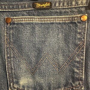 Pode incluir: Close-up do bolso traseiro de cal&ccedil;as de ganga Wrangler azuis. O bolso tem um design costurado em castanho e &eacute; fixado com rebites cor de cobre. O log&oacute;tipo Wrangler &eacute; vis&iacute;vel numa pequena etiqueta acima do bolso.