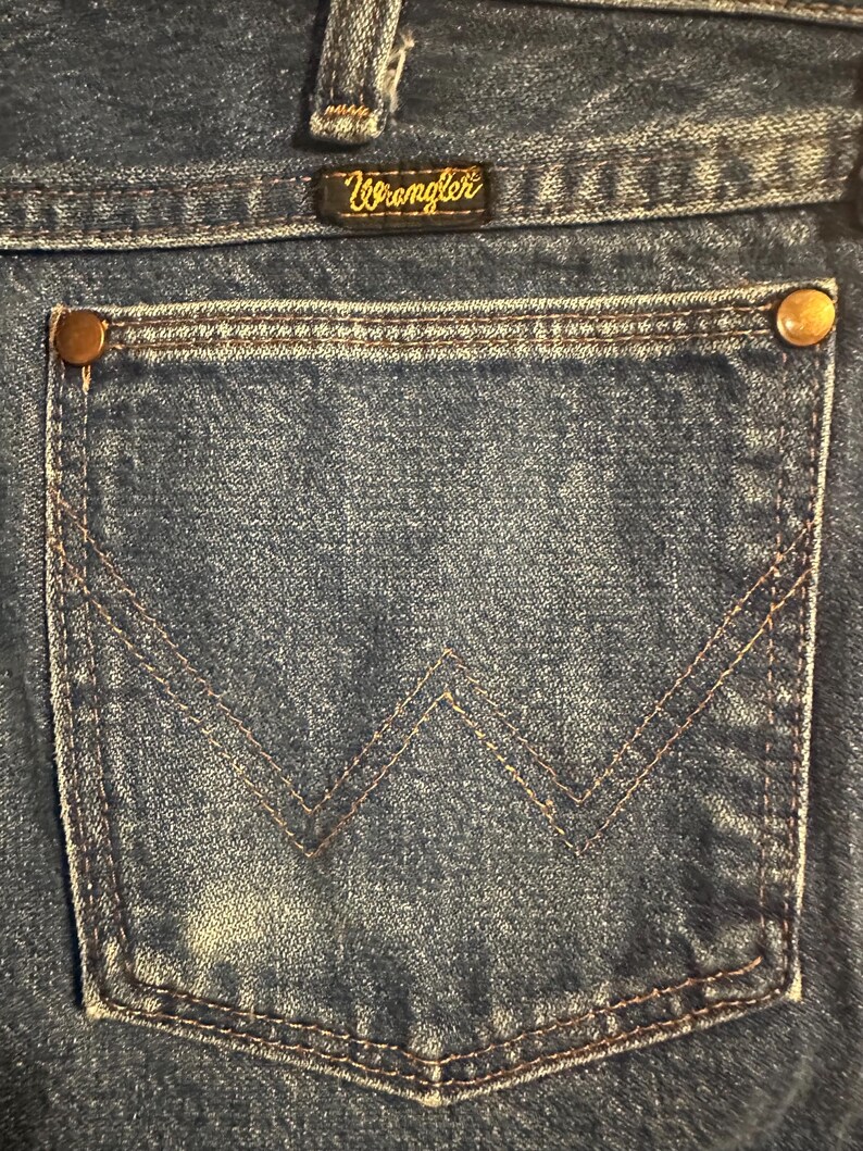 Pode incluir: Close-up do bolso traseiro de cal&ccedil;as de ganga Wrangler azuis. O bolso tem um design costurado em castanho e &eacute; fixado com rebites cor de cobre. O log&oacute;tipo Wrangler &eacute; vis&iacute;vel numa pequena etiqueta acima do bolso.