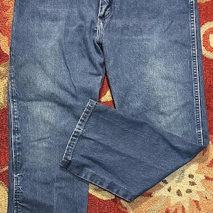 Pode incluir: Cal&ccedil;as de ganga azuis com um design cl&aacute;ssico de cinco bolsos. As cal&ccedil;as de ganga t&ecirc;m um corte de perna reto e um fecho de bot&atilde;o e z&iacute;per. A ganga tem um aspeto desbotado, com &aacute;reas mais claras nas coxas e joelhos. As cal&ccedil;as de ganga s&atilde;o exibidas num tapete estampado.
