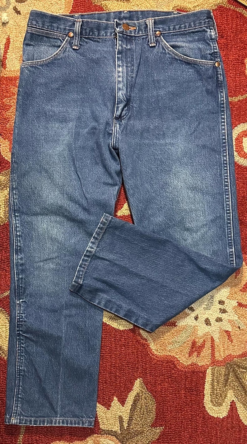 Pode incluir: Cal&ccedil;as de ganga azuis com um design cl&aacute;ssico de cinco bolsos. As cal&ccedil;as de ganga t&ecirc;m um corte de perna reto e um fecho de bot&atilde;o e z&iacute;per. A ganga tem um aspeto desbotado, com &aacute;reas mais claras nas coxas e joelhos. As cal&ccedil;as de ganga s&atilde;o exibidas num tapete estampado.