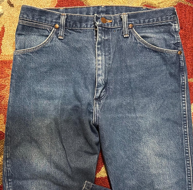 Pode incluir: Close-up de jeans azuis. Os jeans t&ecirc;m um design cl&aacute;ssico de cinco bolsos com fecho de bot&atilde;o e z&iacute;per. O denim tem uma apar&ecirc;ncia desbotada, com desgaste vis&iacute;vel. Os jeans s&atilde;o provavelmente vintage ou usados.